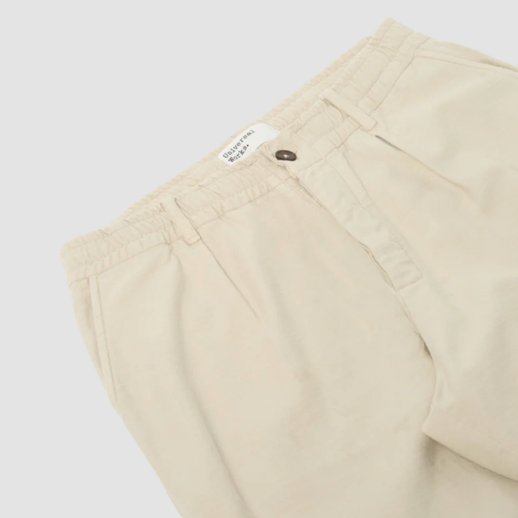 Universal Works Pleated Track Pant in Driftwood Slub Sateen: Loose-fit, tapered leg pant made of 100% cotton. Perfect for summer with its unique texture. Mid-rise with front and rear pockets