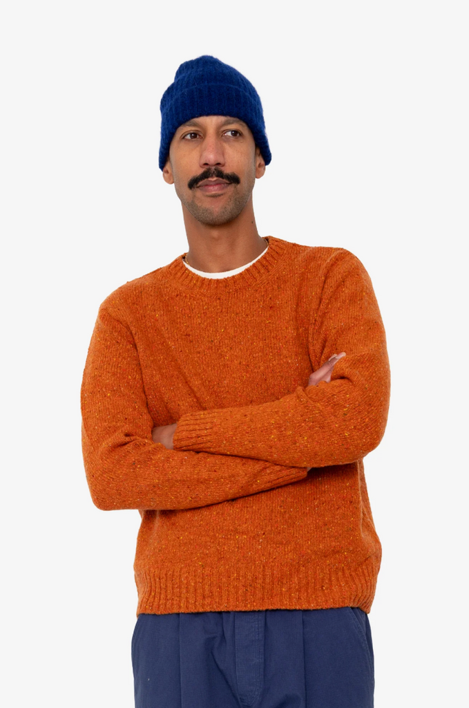 Man wearing an orange sweater and blue beanie with arms crossed on a white background.