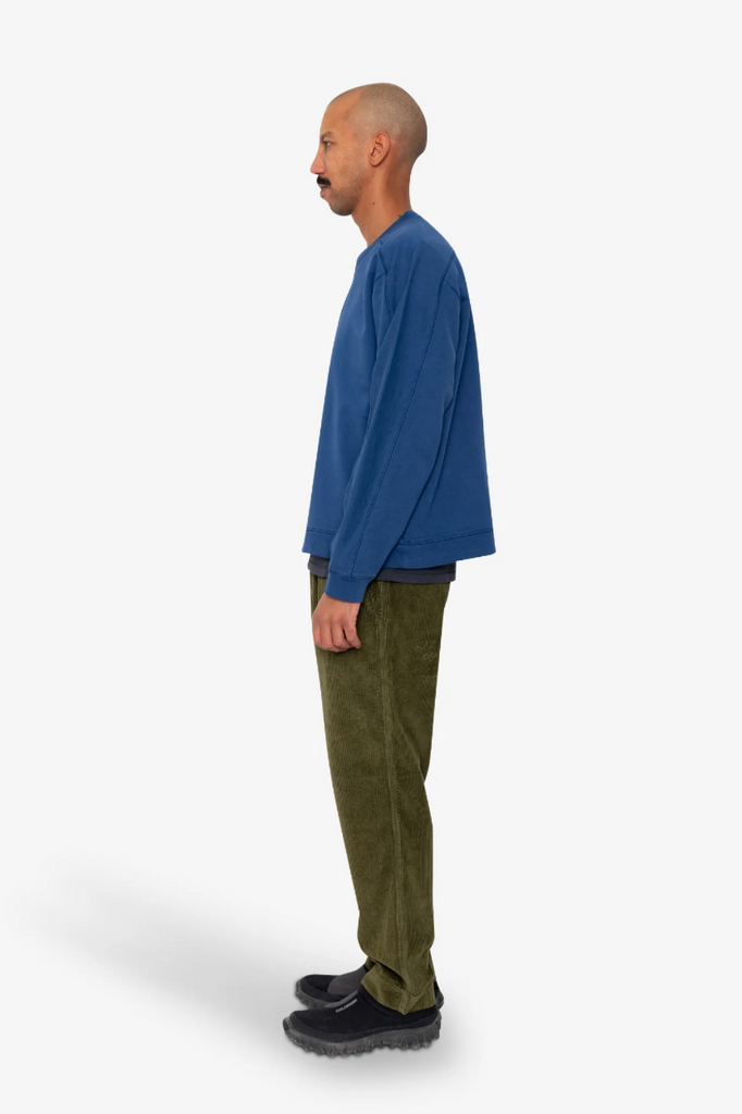 Man wearing a blue sweater and green pants standing against a white background.