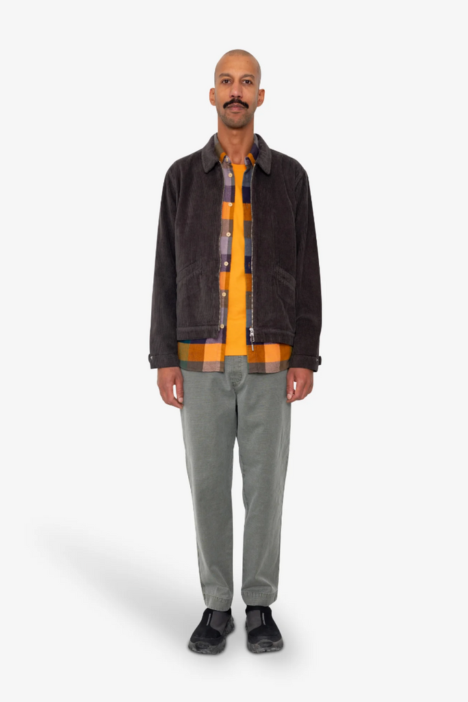 Man wearing a dark corduroy jacket over a plaid shirt with a yellow pattern, gray pants, and black shoes on a white background.