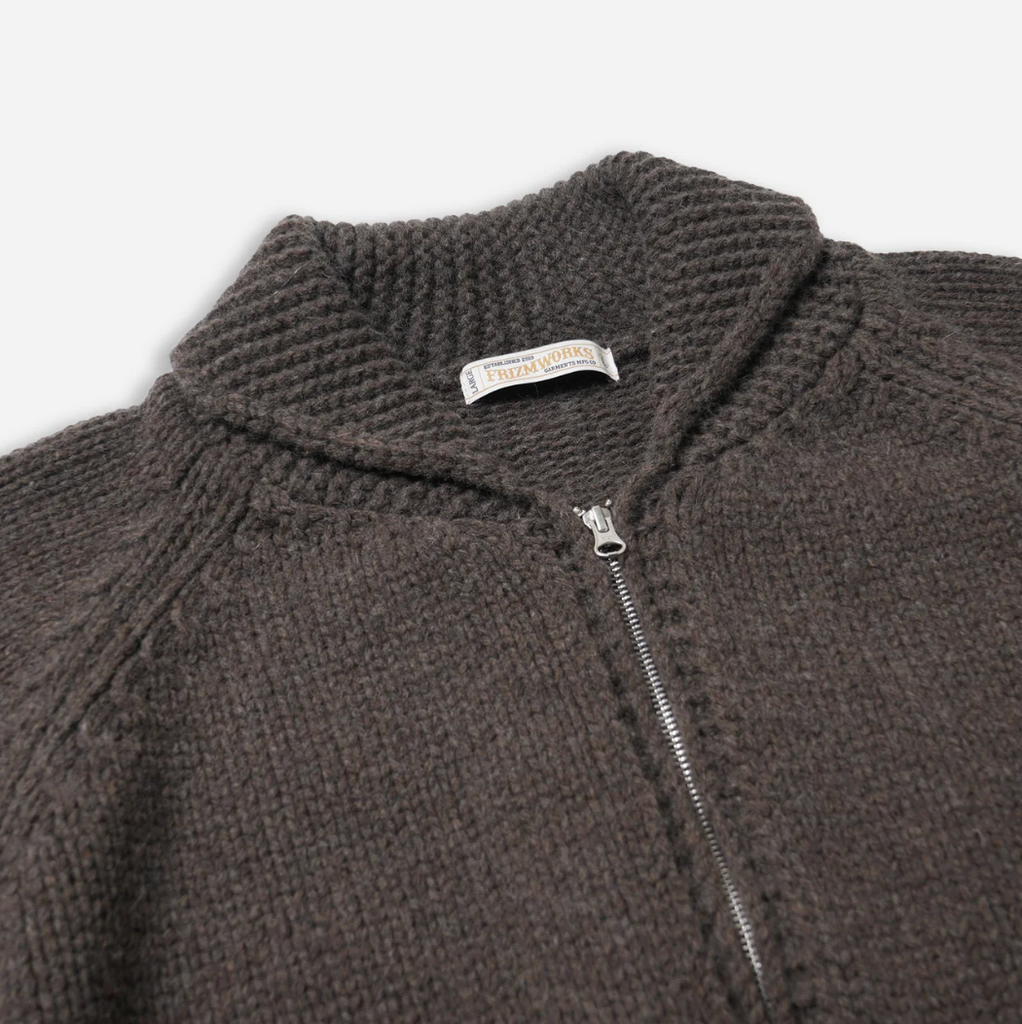 Close-up of a brown knit sweater with a zipper, featuring a brand label on a light gray background.