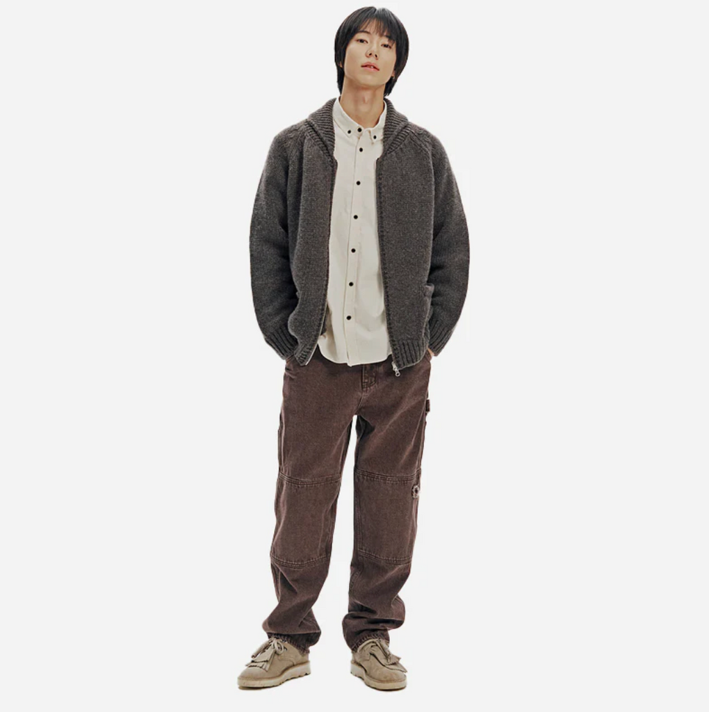 Person wearing a brown knitted zip up cardigan over a white shirt and brown pants on a white background.