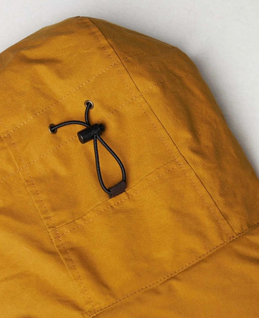 Close up of a Yellow wax jacket with hood on a light grey background.