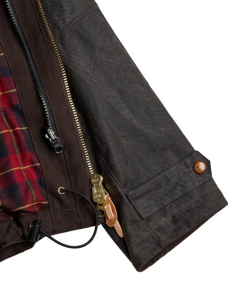 Close up of a brown wax jacket with zip fastenings on a light grey background.