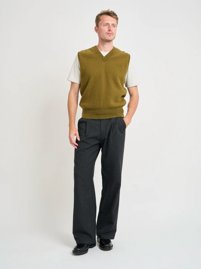 Man wearing a green sweater vest over a white shirt with black pants on a white background.