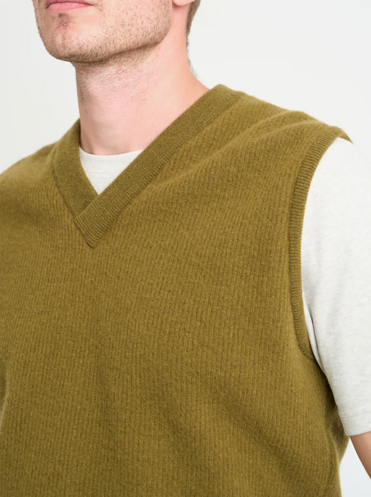 Man wearing a green knitted vest over a white shirt on a plain background.