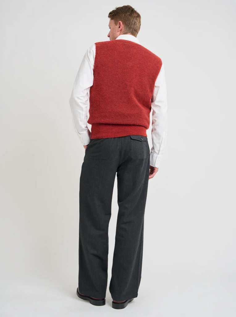 Man wearing a red sweater vest over a white shirt with gray pants on a white background.