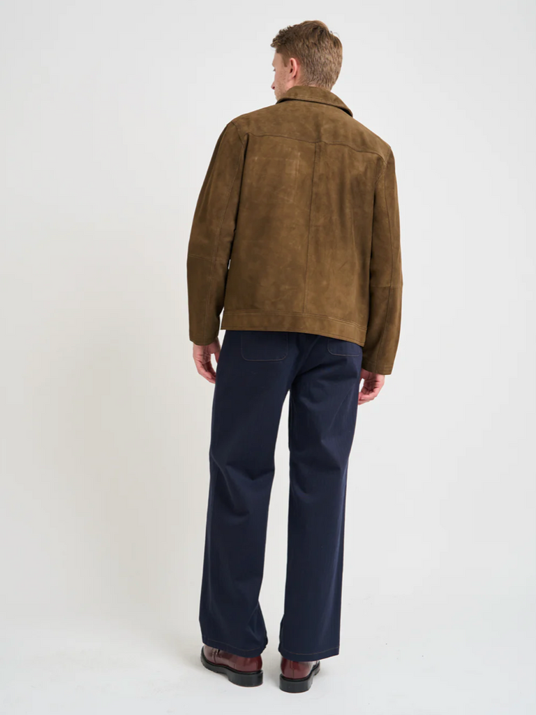Man wearing a brown suede jacket and dark blue pants on a white background.