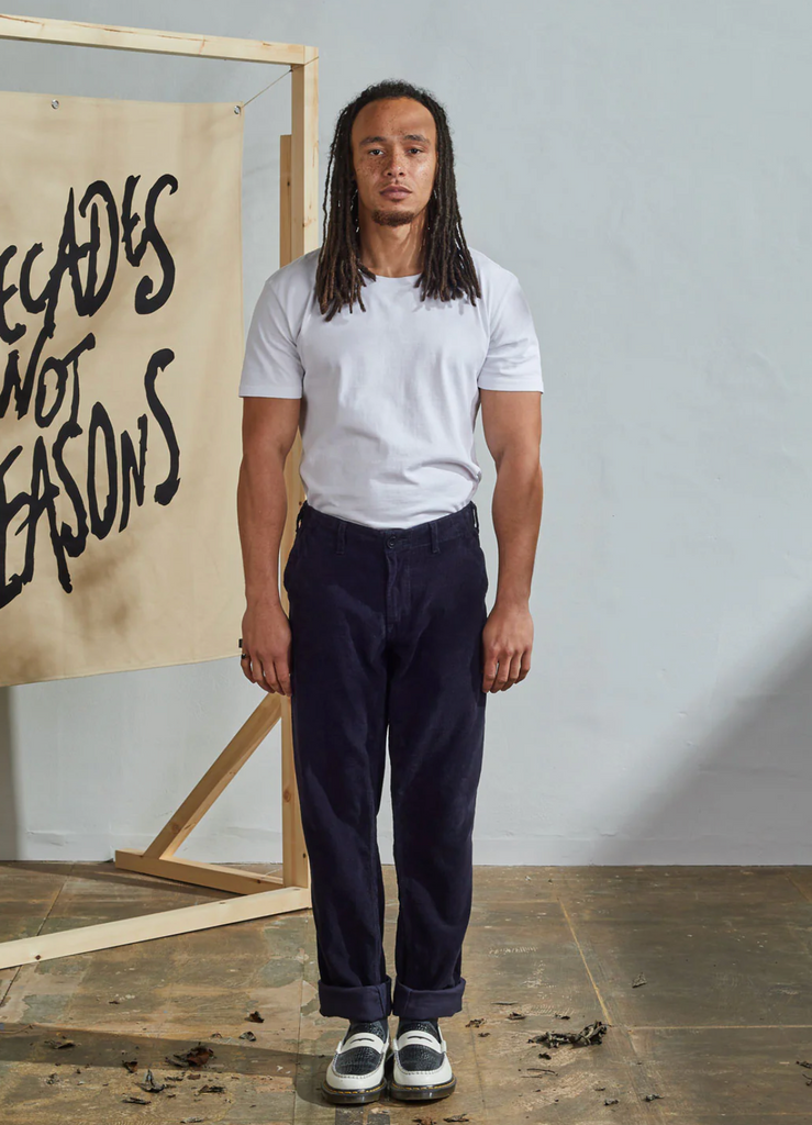 Person wearing a white t-shirt and dark pants standing in front of a wall with text.