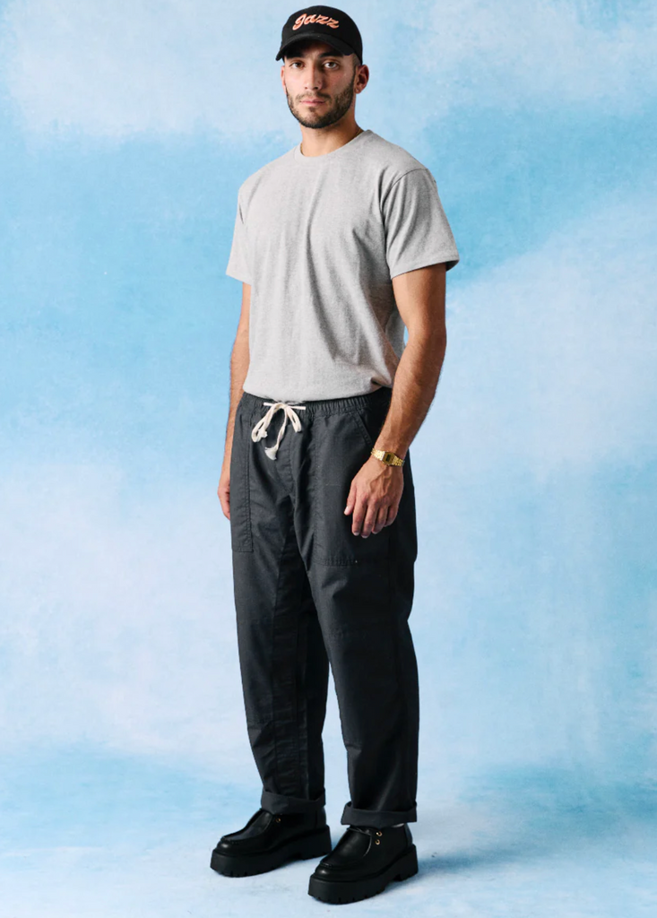 Man wearing a gray t-shirt, black pants, and a black cap against a light blue gradient background.