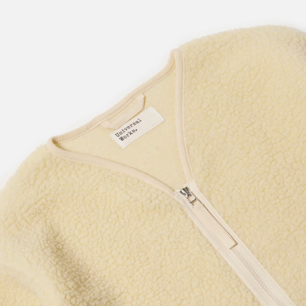 Close-up of an ecru fleece jacket with a zipper and 'Universal Works' label on a white background..
