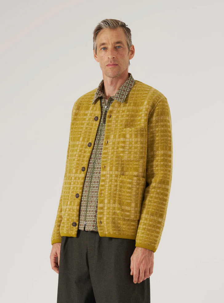 Man wearing a mustard yellow textured jacket over a patterned shirt on a plain background.